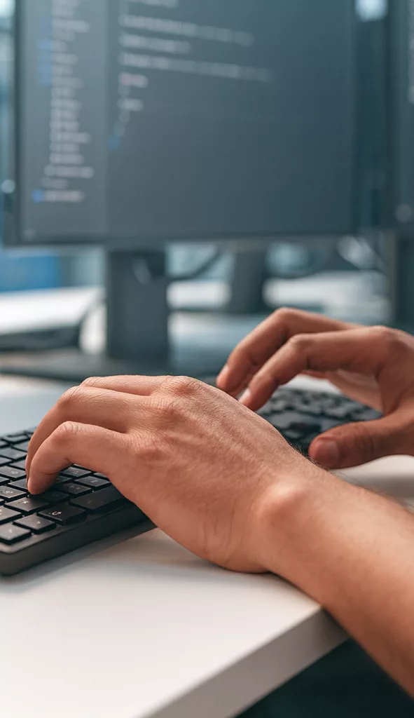 Nahaufnahme von Händen an der Tastatur, im Hintergrund Code auf einem Bildschirm